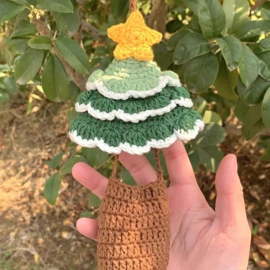 A crochet Christmas tree with a yellow star on top, made from green and white yarn, hanging in front of green plants and trees.